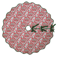 A round Christmas tree skirt with a scalloped edge, featuring a red background with a white and green pine tree and snowflake pattern, finished with dark green ribbon ties.