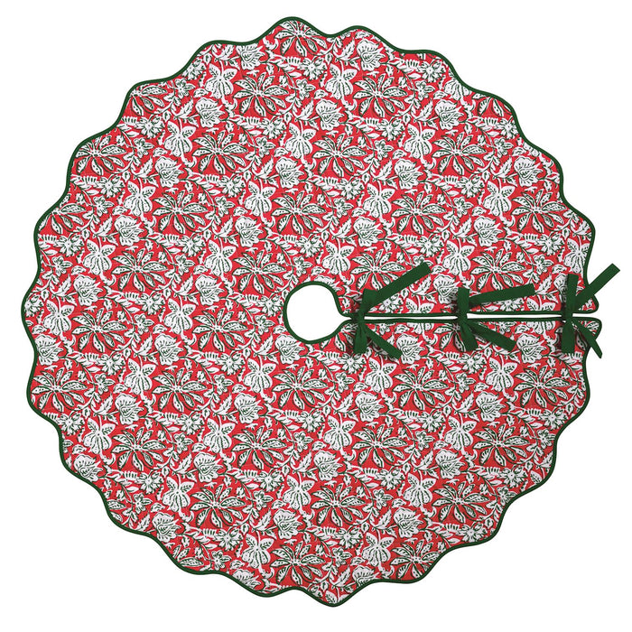 A round Christmas tree skirt with a scalloped edge, featuring a red background with a white and green pine tree and snowflake pattern, finished with dark green ribbon ties.