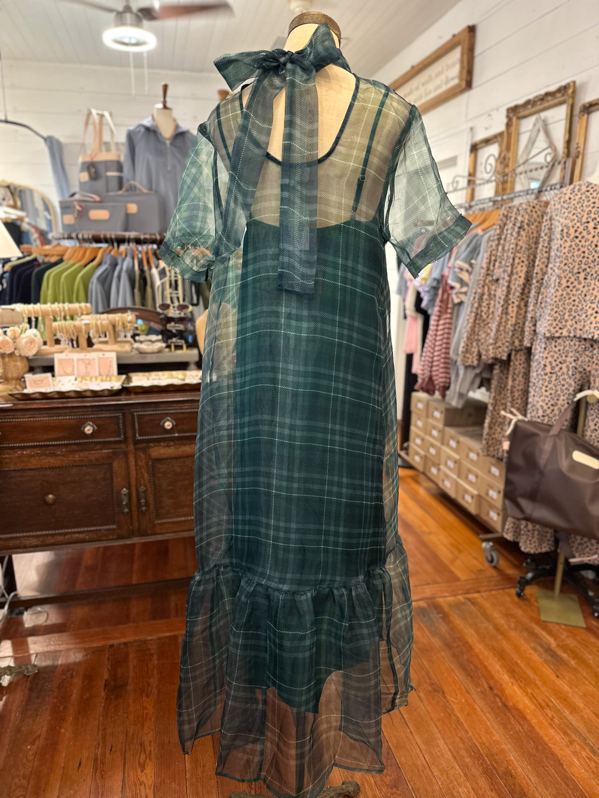 A dark green plaid midi dress displayed on a mannequin in a clothing boutique. The dress features a sheer mesh overlay with a matching bow at the back neckline, short sleeves, and a ruffled hemline.