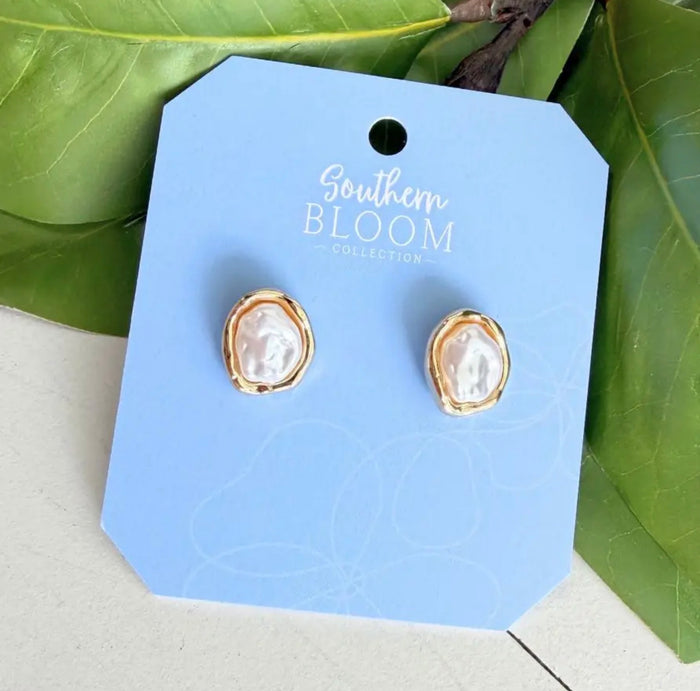 A pair of Southern Bloom Collection stud earrings with irregular white baroque pearls in gold bezel settings, displayed on a light blue branded card with green leaves in the background.