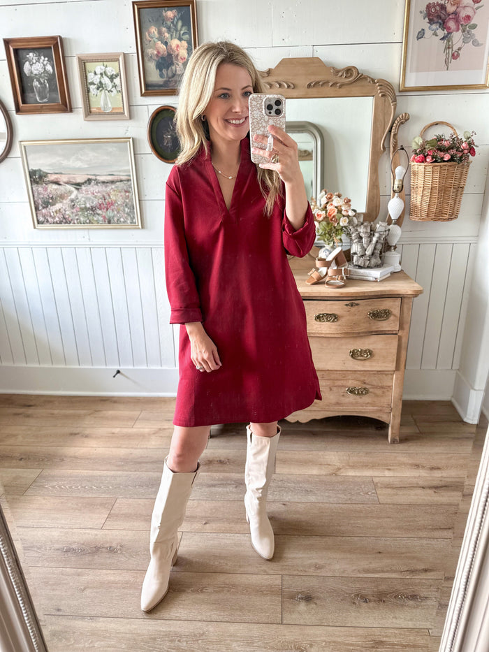 Woman in a red dress and white knee-high boots taking a mirror selfie in a room with wooden floor and dresser.