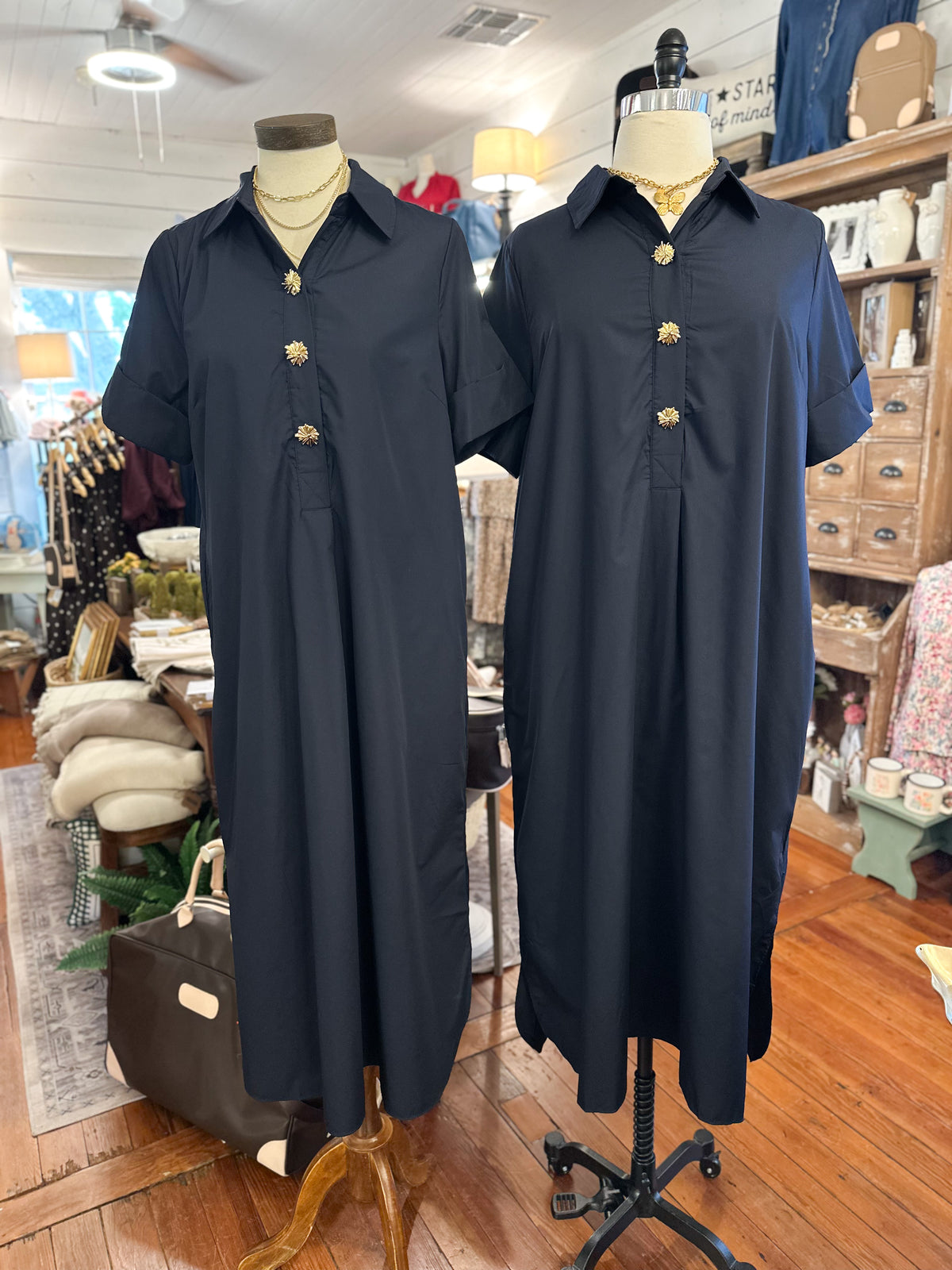 Two navy blue shift dresses displayed on dress forms in a boutique setting. The dresses feature short sleeves, a button-down collar, and a front button placket with decorative gold button details.