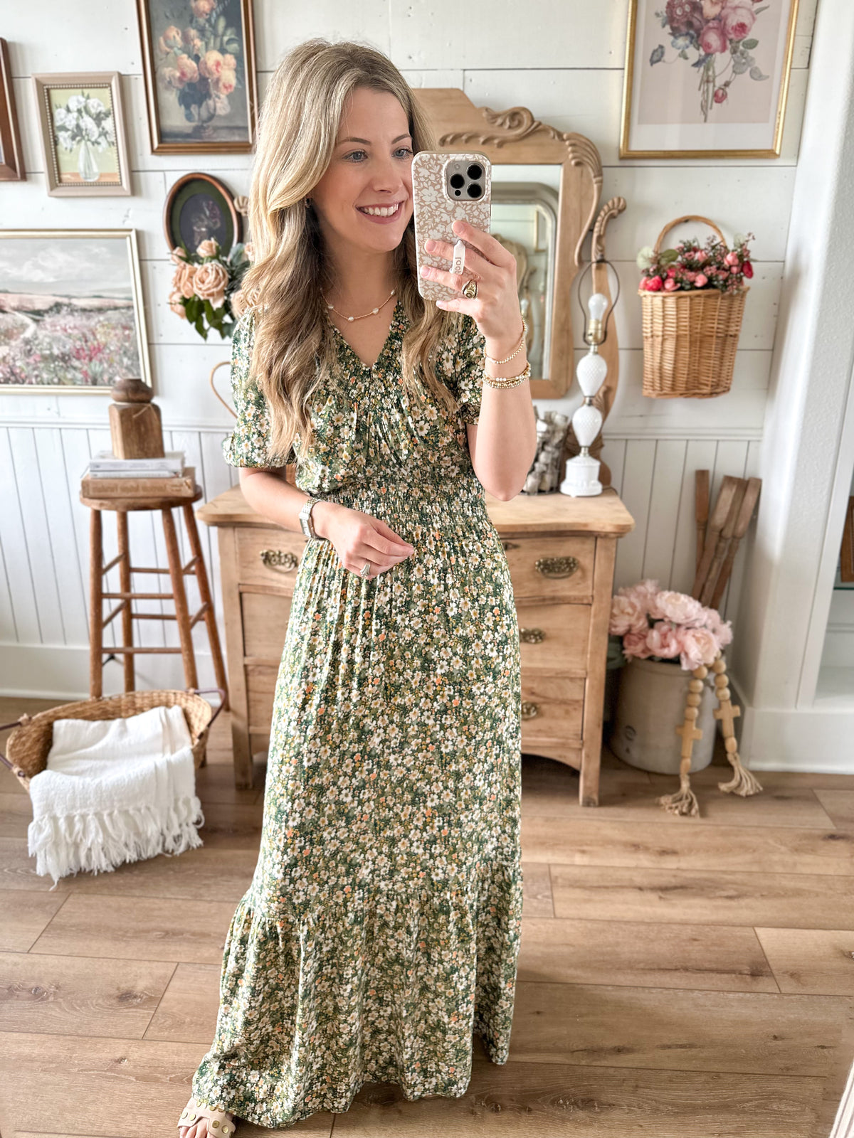 Woman in a floral dress taking a mirror selfie in a home setting with wooden furniture and decor.