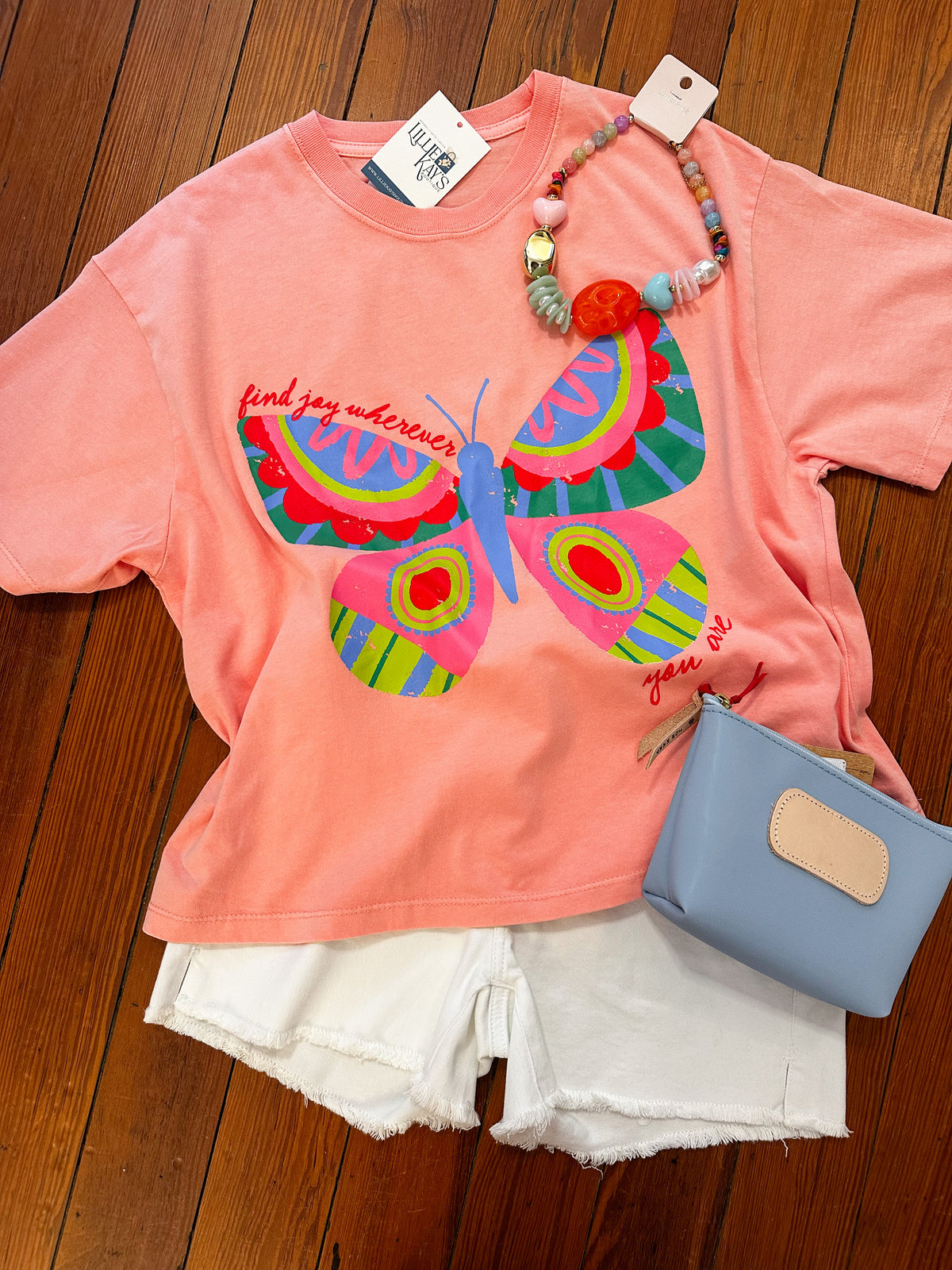 Pink t-shirt with colorful butterfly design, white shorts, and blue pouch on a wooden surface