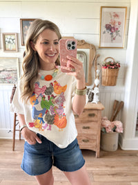 Woman taking a mirror selfie wearing a colorful graphic t-shirt and denim shorts in a home setting.