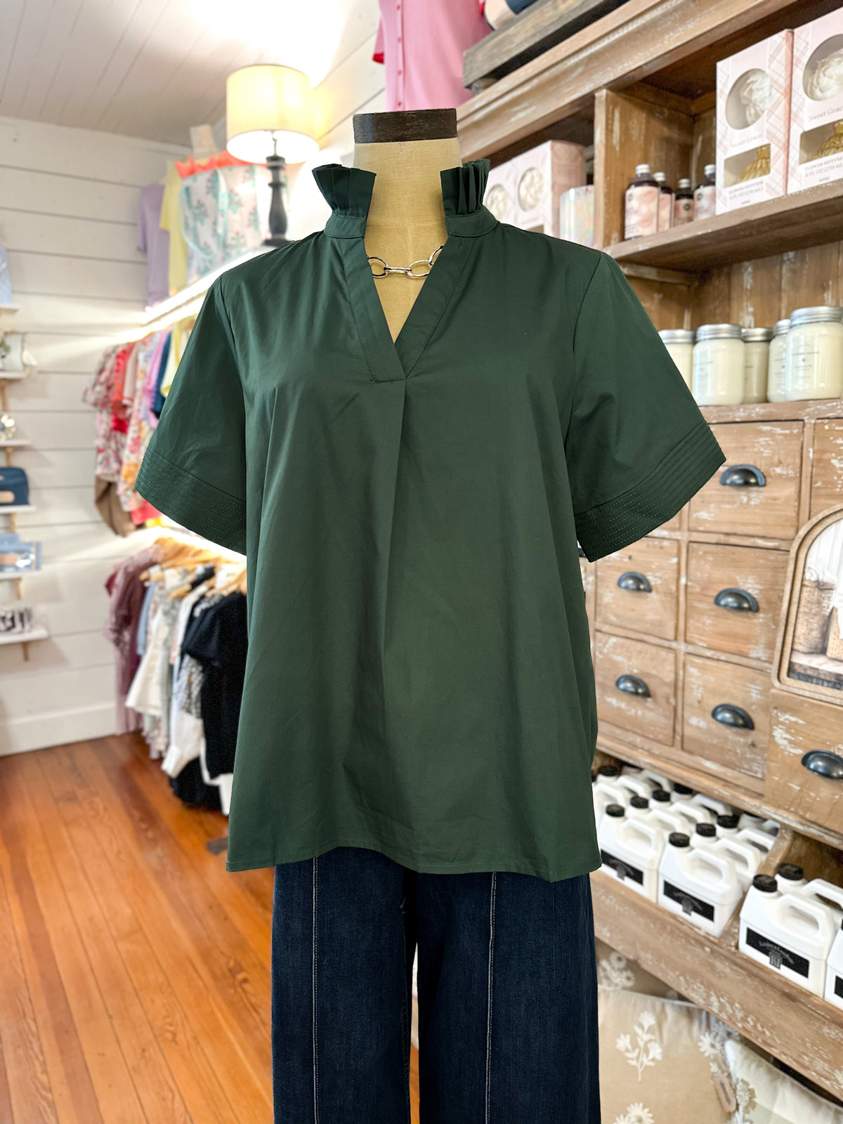 A dark green short-sleeve blouse with a split V-neckline displayed on a mannequin torso inside a retail store. The blouse is styled with dark blue wide-leg pants, with wooden shelving and clothing racks visible in the background.