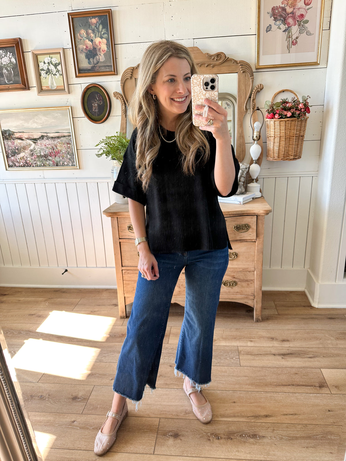 Woman taking a mirror selfie in a room with wooden floor and framed pictures on the wall. black short sleeve sweater top 