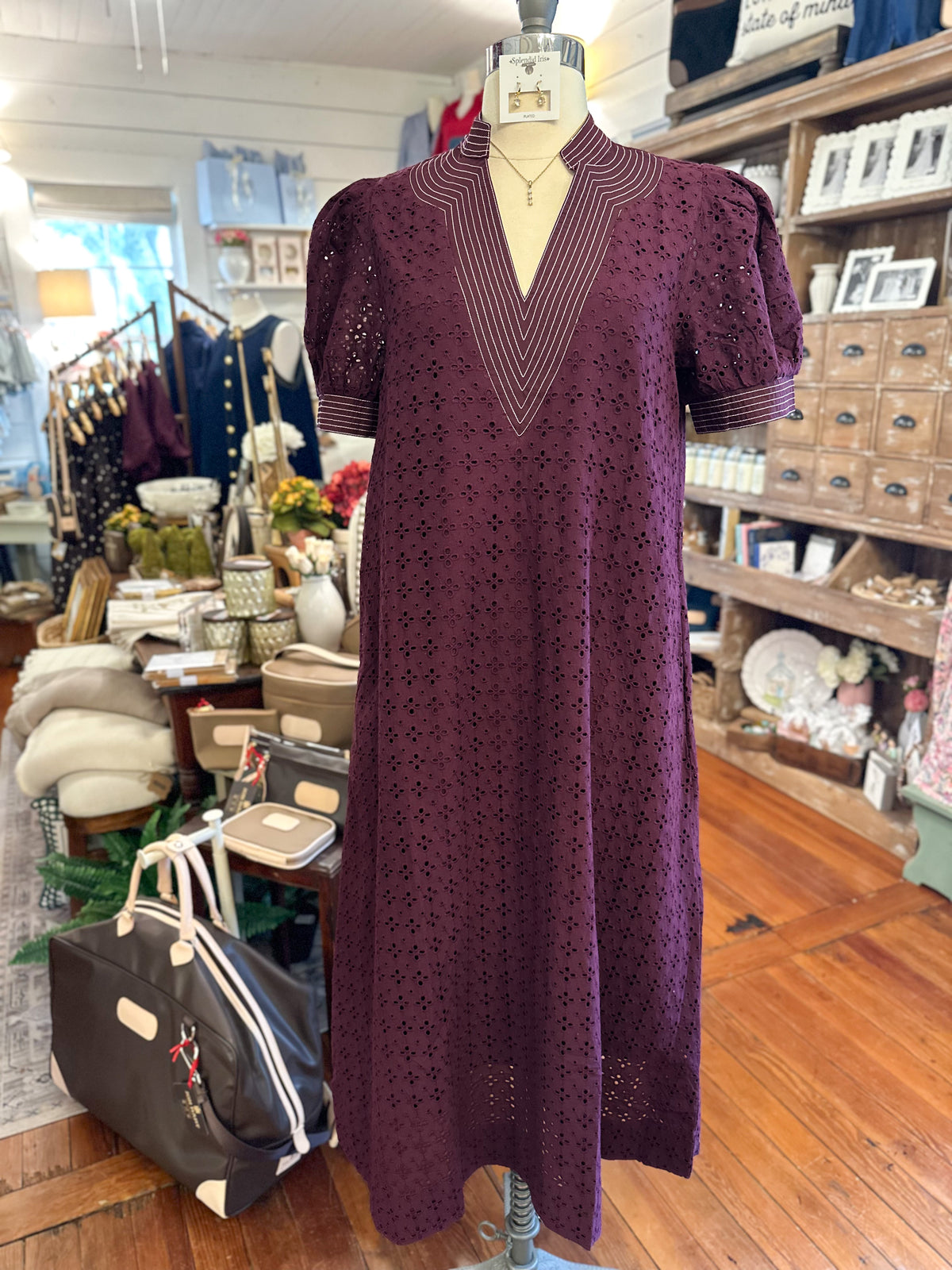 A dark purple eyelet midi dress with puff sleeves and a V-neckline, displayed on a mannequin inside a clothing boutique.
