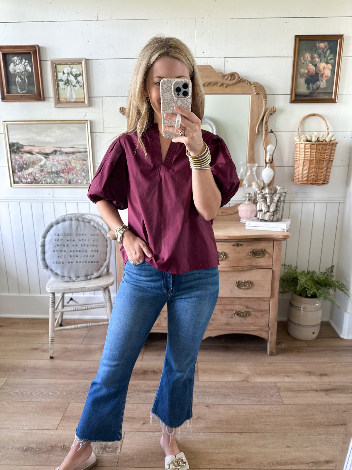 Person taking a mirror selfie wearing a burgundy shirt and blue jeans in a room with wooden floor and white walls.