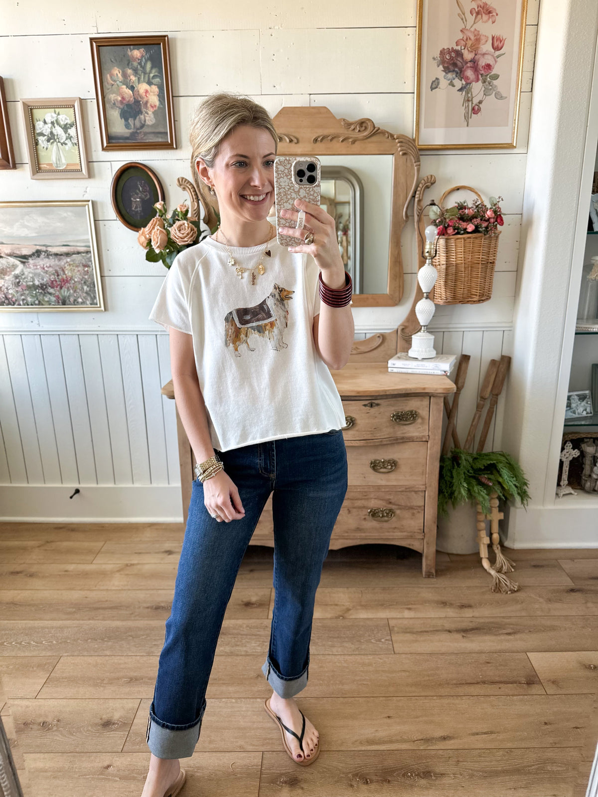 Woman taking a mirror selfie in a room with wooden floors and framed pictures on the wall.