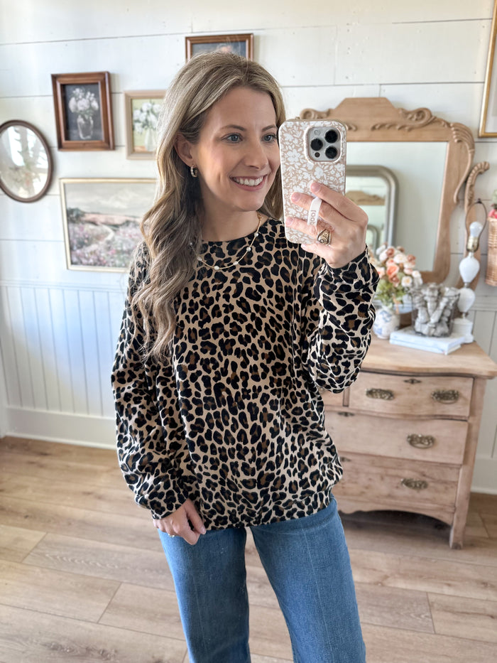 Woman taking a mirror selfie wearing a leopard print sweater and blue jeans in a room with wooden furniture and decor.