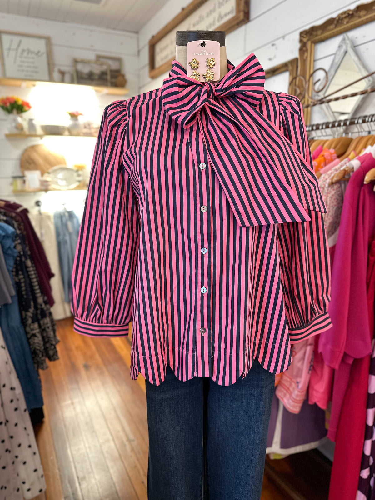 A pink and pine green vertically striped blouse with a large matching bow at the collar, displayed on a mannequin in a clothing boutique.