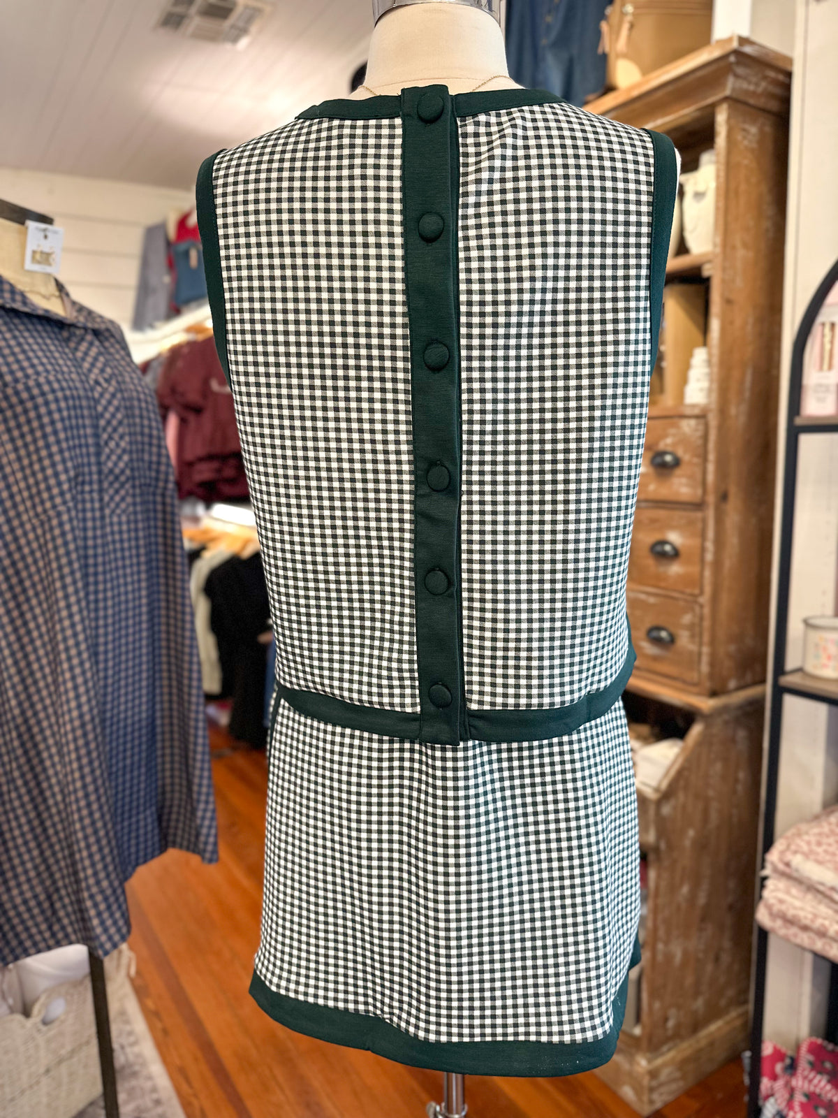 A sleeveless green and white gingham apron-style vest with dark green trim and a vertical button placket, displayed on a mannequin in a clothing store.