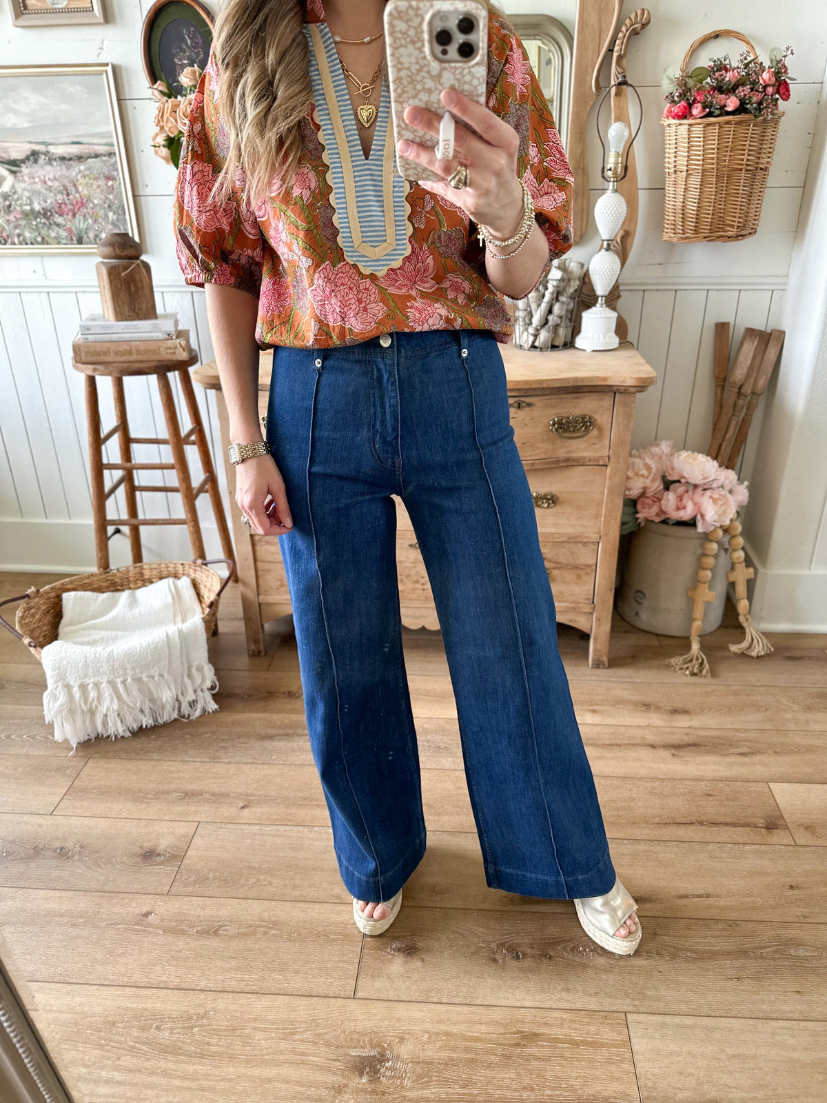 Person taking a mirror selfie wearing a floral blouse and blue jeans in a home setting.