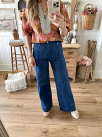 Person taking a mirror selfie wearing a floral blouse and blue jeans in a home setting.