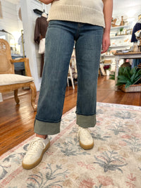 Person wearing blue jeans and white sneakers on a patterned rug indoors.