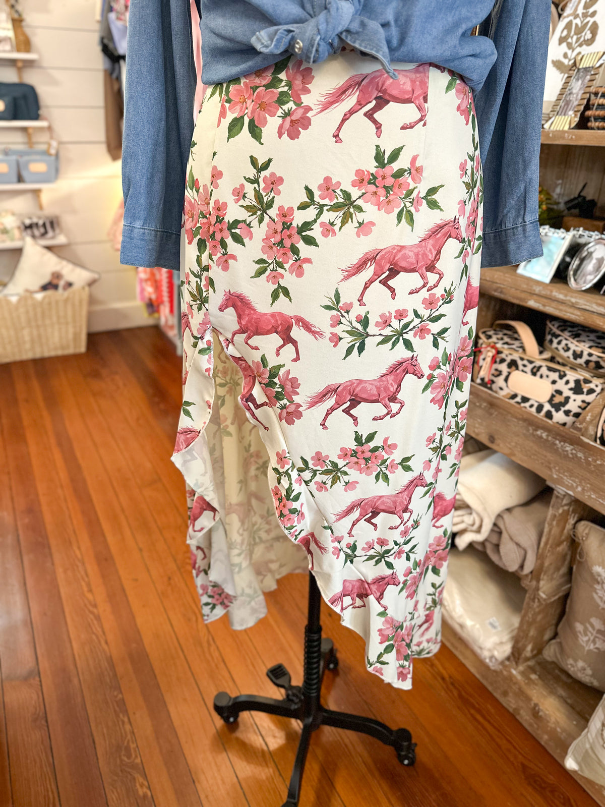 A floral print midi skirt with pink horses and pink flowers on a white background, styled with a blue denim shirt tied at the waist on a mannequin in a retail store.