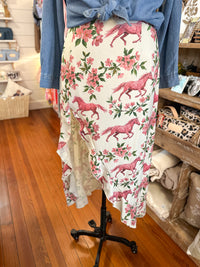 A floral print midi skirt with pink horses and pink flowers on a white background, styled with a blue denim shirt tied at the waist on a mannequin in a retail store.