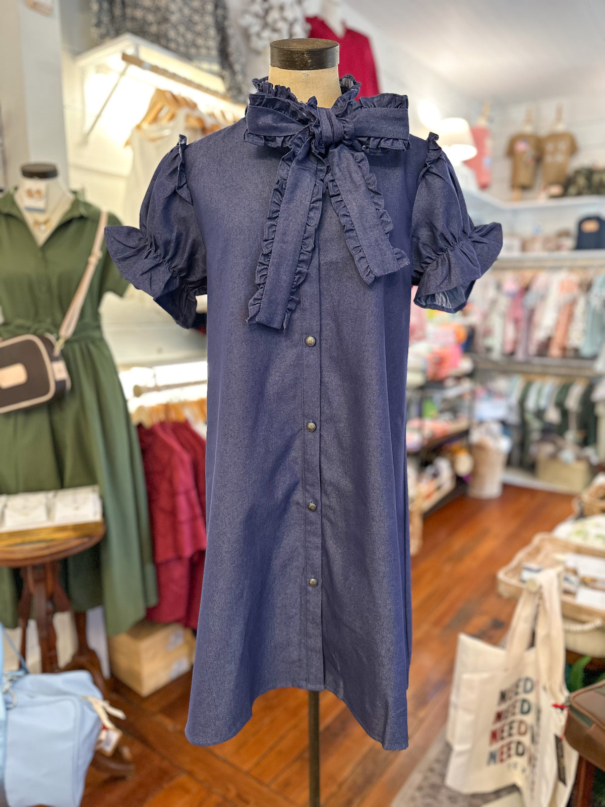 A blue denim-like button-front dress with ruffled trim and ruffled short sleeves, displayed on a mannequin inside a clothing boutique.