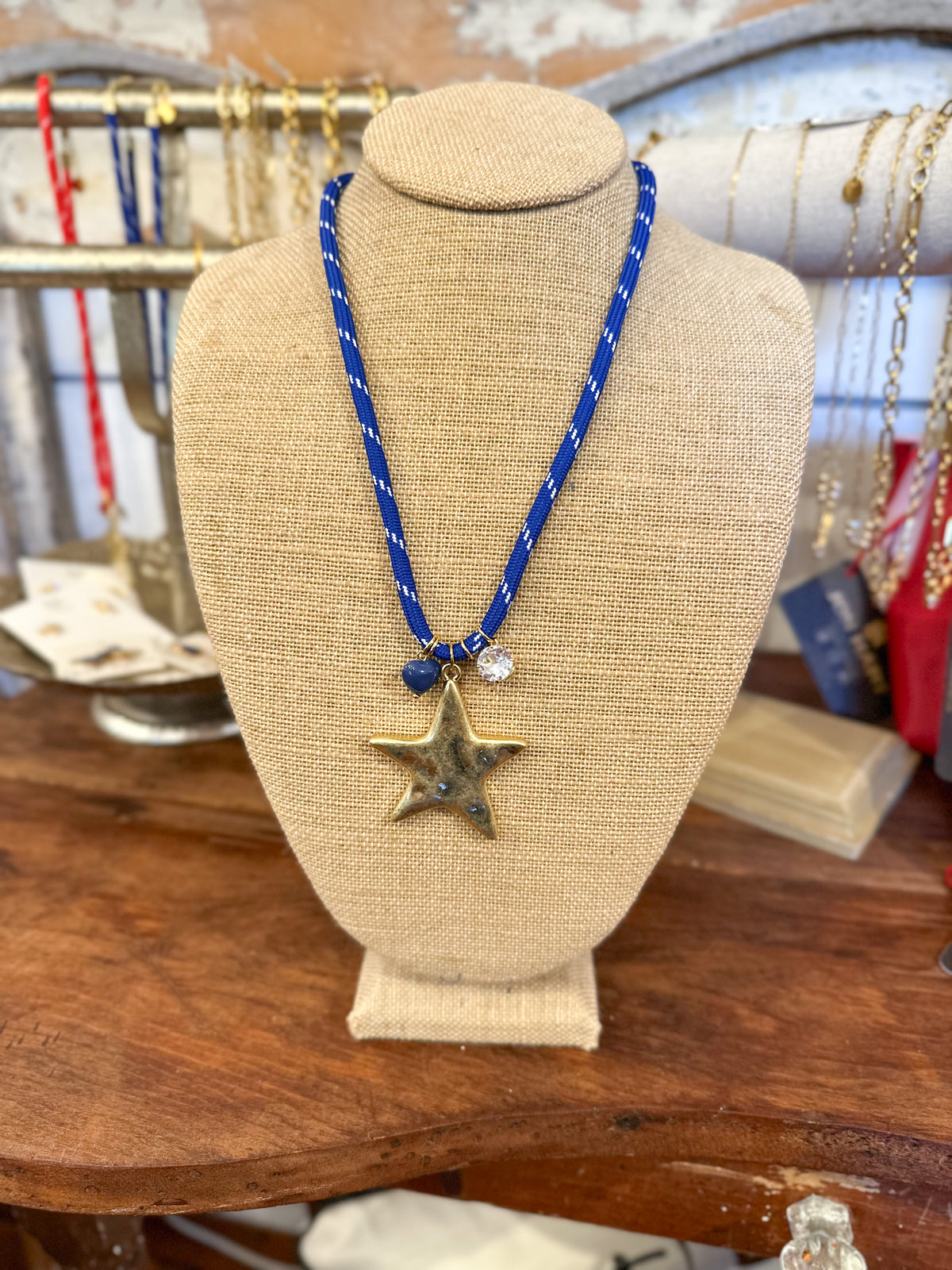 A blue braided cord necklace with a bronze star pendant and a small clear bead charm, displayed on a beige fabric jewelry bust on a wooden table.