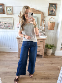 Woman taking a mirror selfie wearing a gray sweater and blue jeans in a room with wooden flooring and white walls.