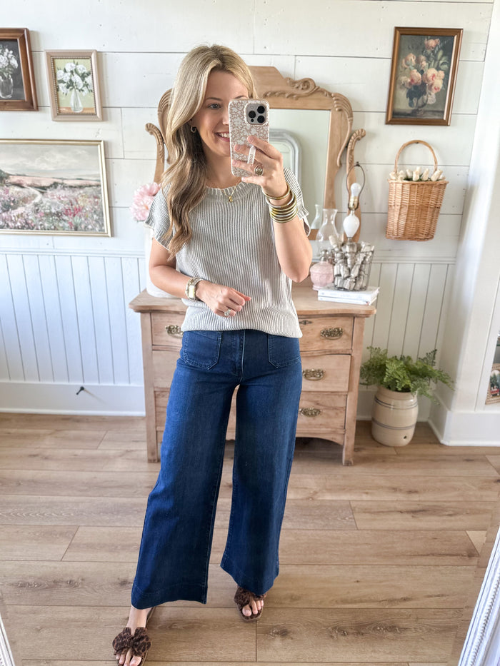 Woman taking a mirror selfie wearing a gray sweater and blue jeans in a room with wooden flooring and white walls.