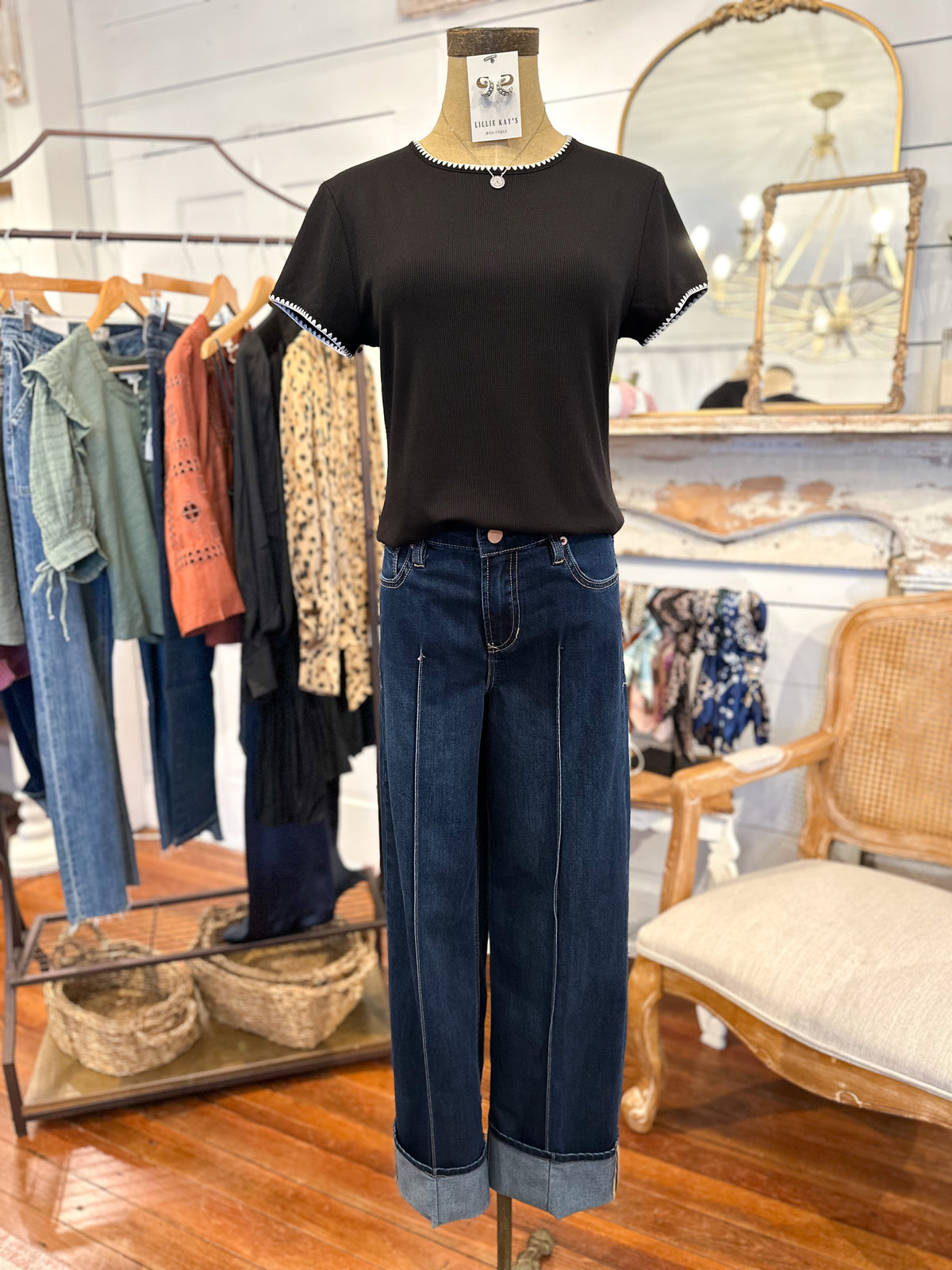 Mannequin wearing a black t-shirt and blue jeans in a store setting with clothing racks and a chair.