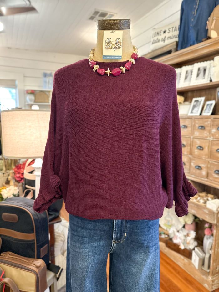 A burgundy knit sweater with three-quarter balloon sleeves displayed on a mannequin torso, styled with blue jeans and a chunky pink beaded necklace in a boutique setting.