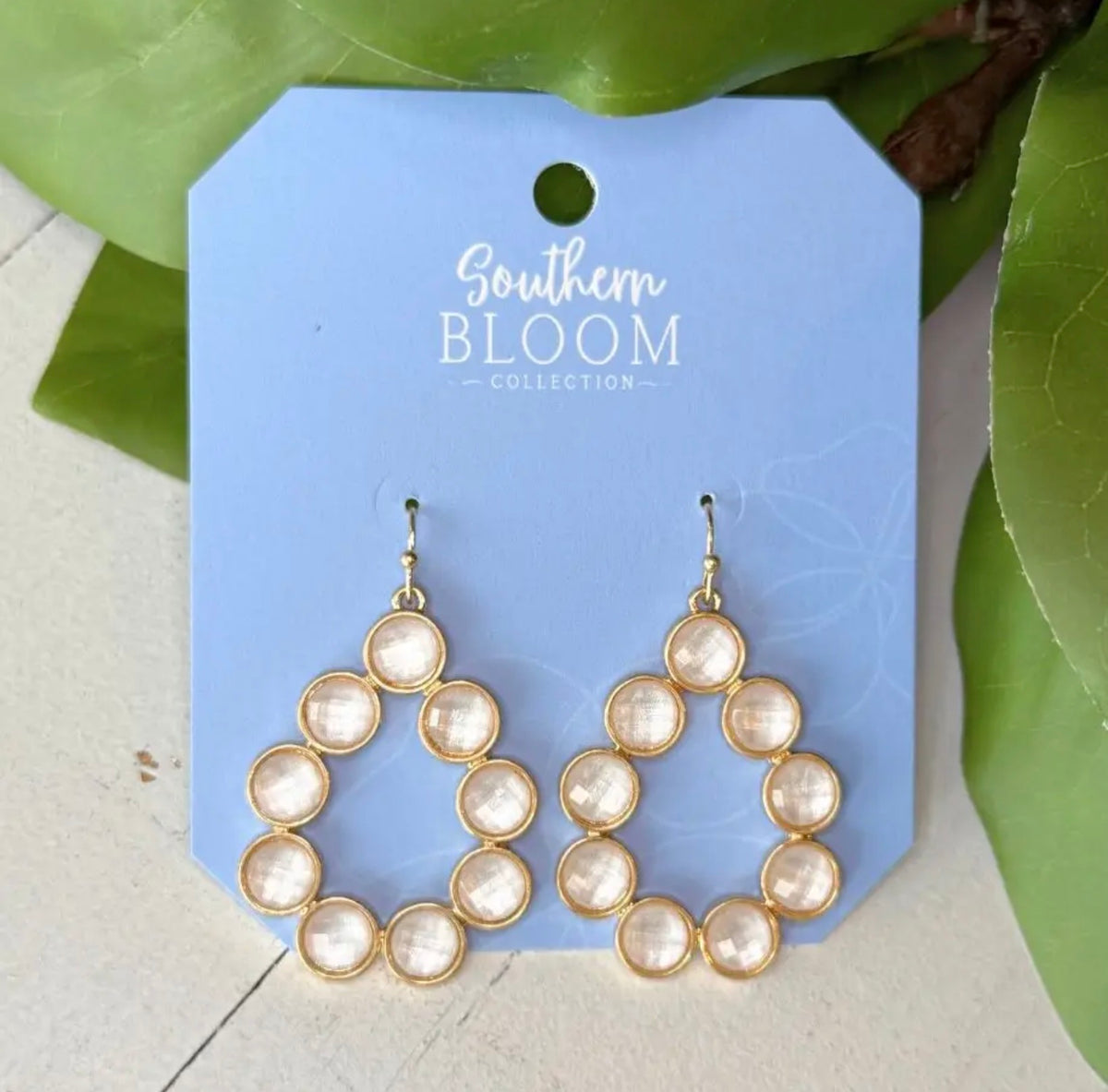 A pair of gold-tone drop earrings with oval clear stones, displayed on a light blue card labeled Southern Bloom Collection, with green leaves in the background.