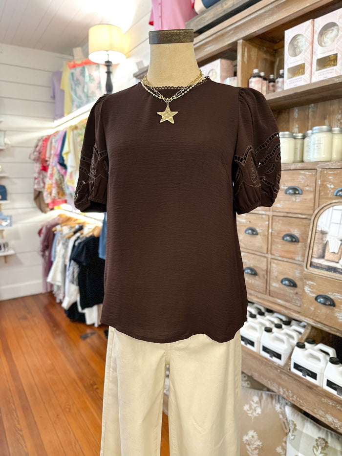 A dark brown short-sleeve blouse with lace-up puff sleeves and a gold star pendant necklace, displayed on a mannequin in a clothing boutique alongside cream pants.