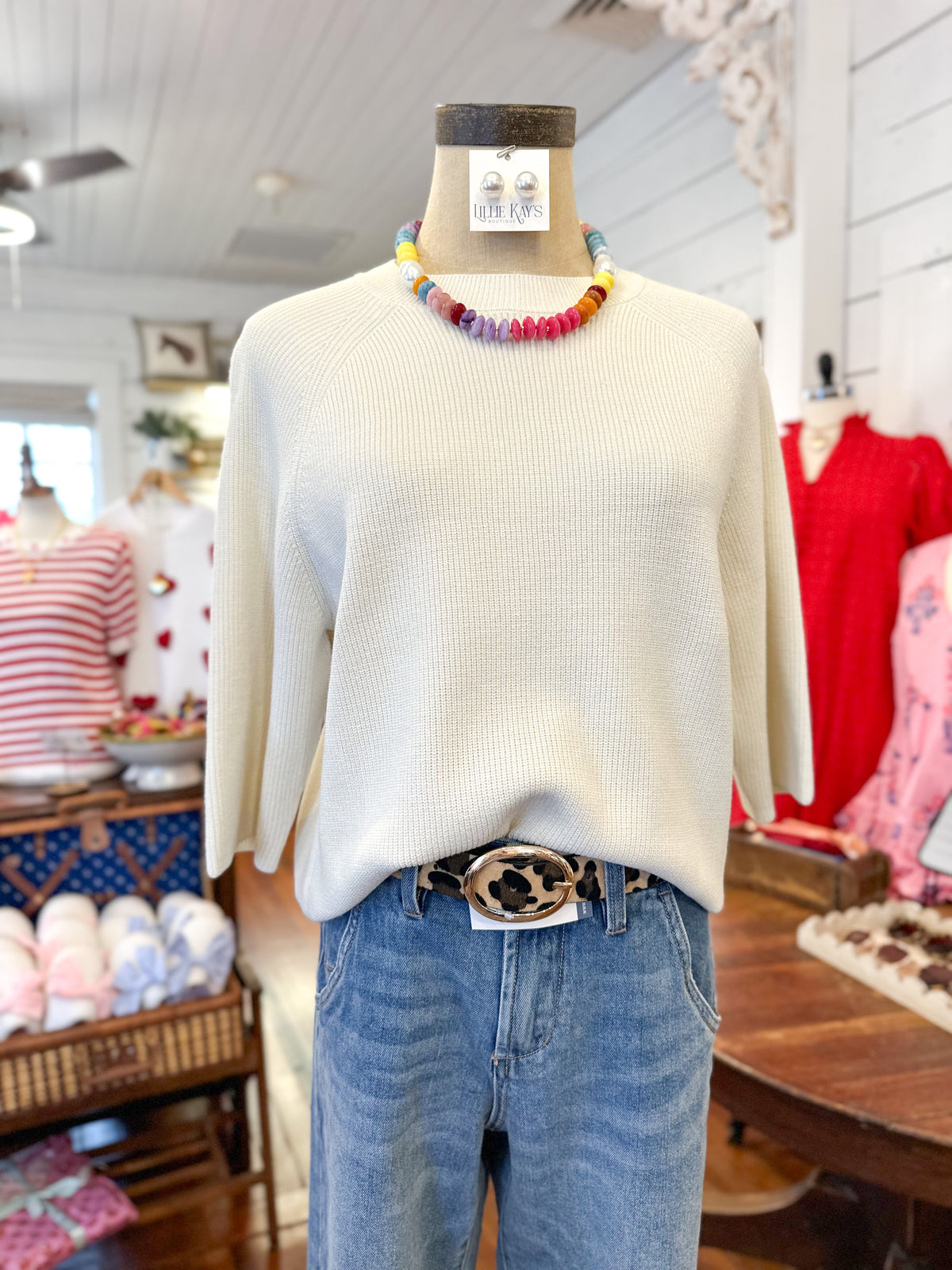 Mannequin wearing a cream sweater and blue jeans with a colorful necklace in a store setting.