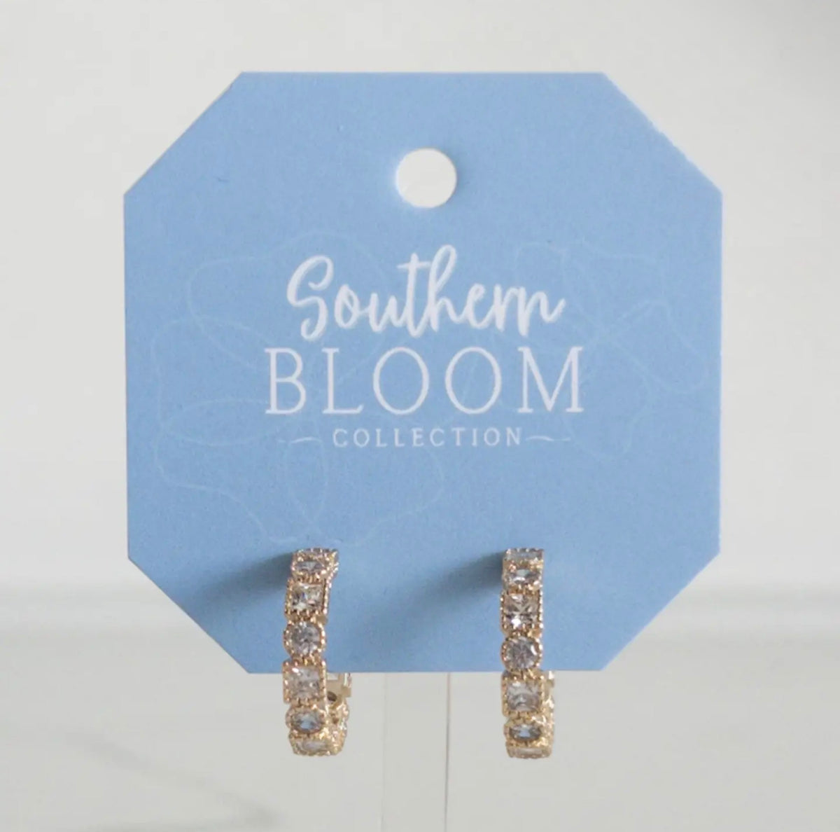 A pair of gold huggie hoop earrings with clear pavé-set stones, displayed on a light blue octagonal card labeled 'Southern BLOOM COLLECTION'.