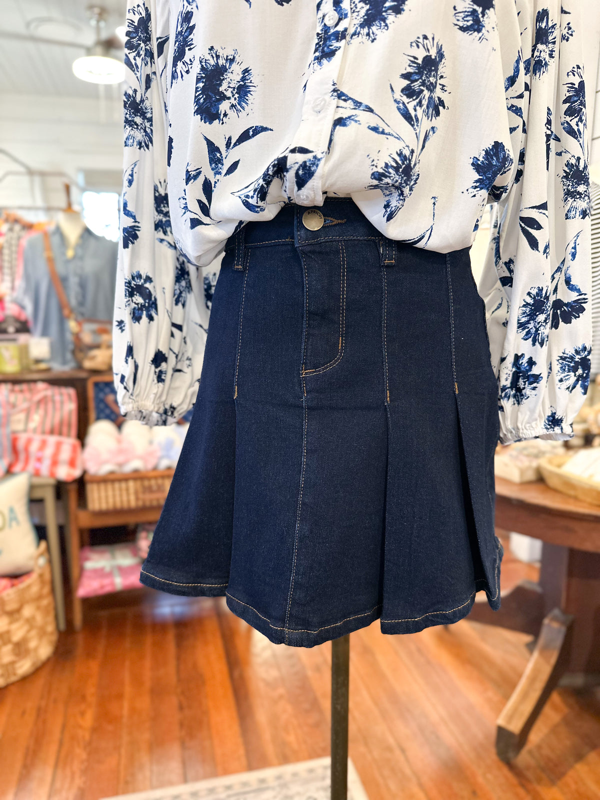 Mannequin wearing a white floral blouse and dark denim pleated skirt in a store setting.