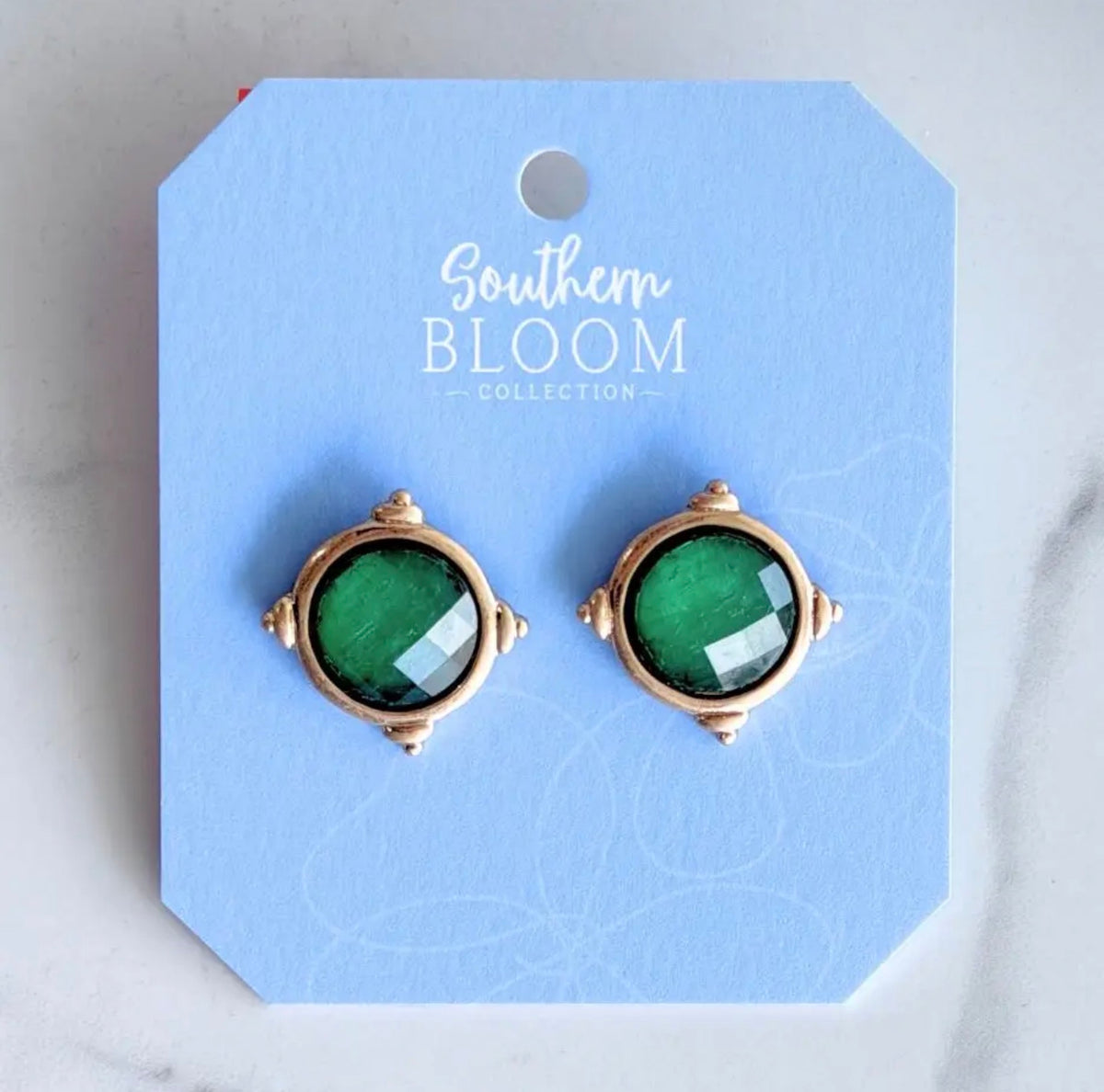 A pair of green  stud earrings in a gold-toned setting, displayed on a light blue card from the Southern Bloom Collection.