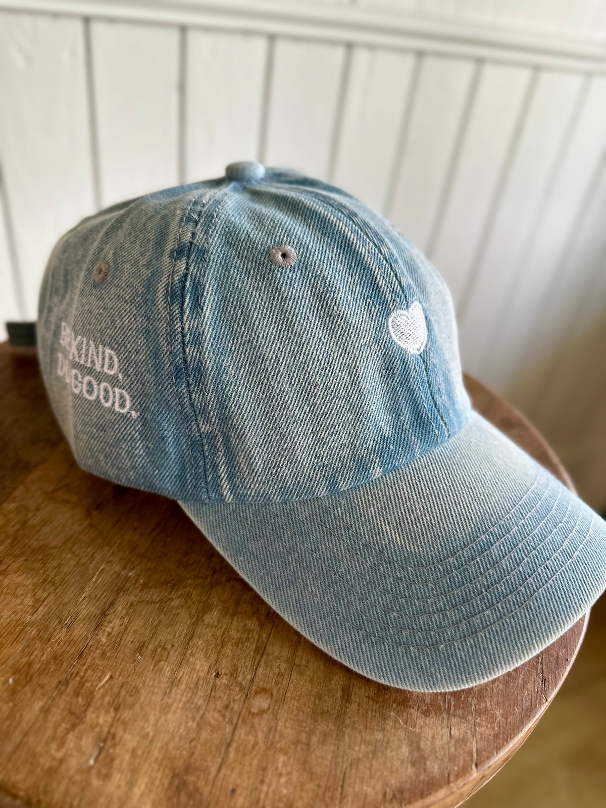 Blue denim cap with 'Be Kind, Be Good' text on a wooden surface.