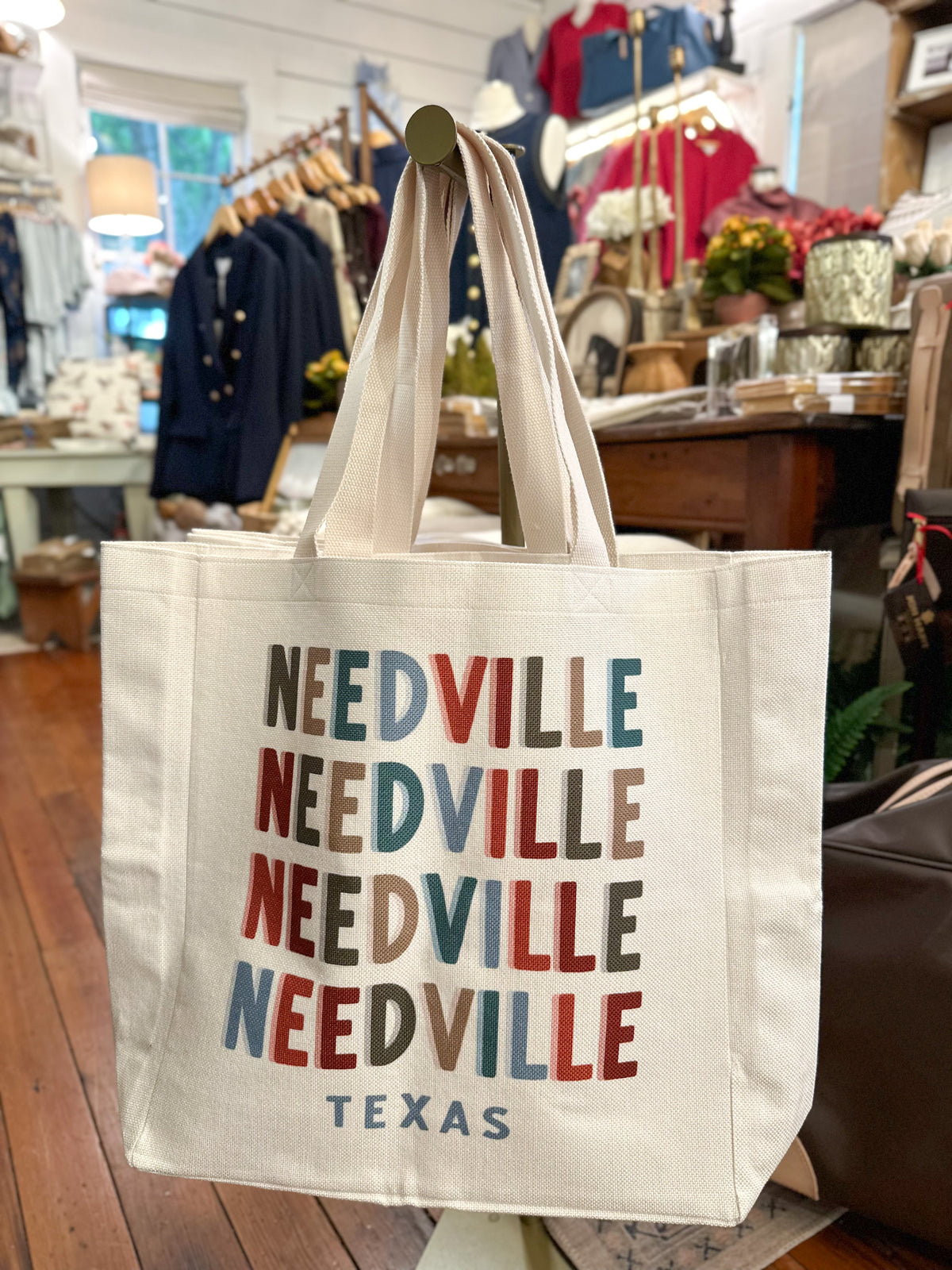 A cream canvas tote bag with 'NEEDVILLE TEXAS' printed in stacked multicolor letters, hanging in a retail store.