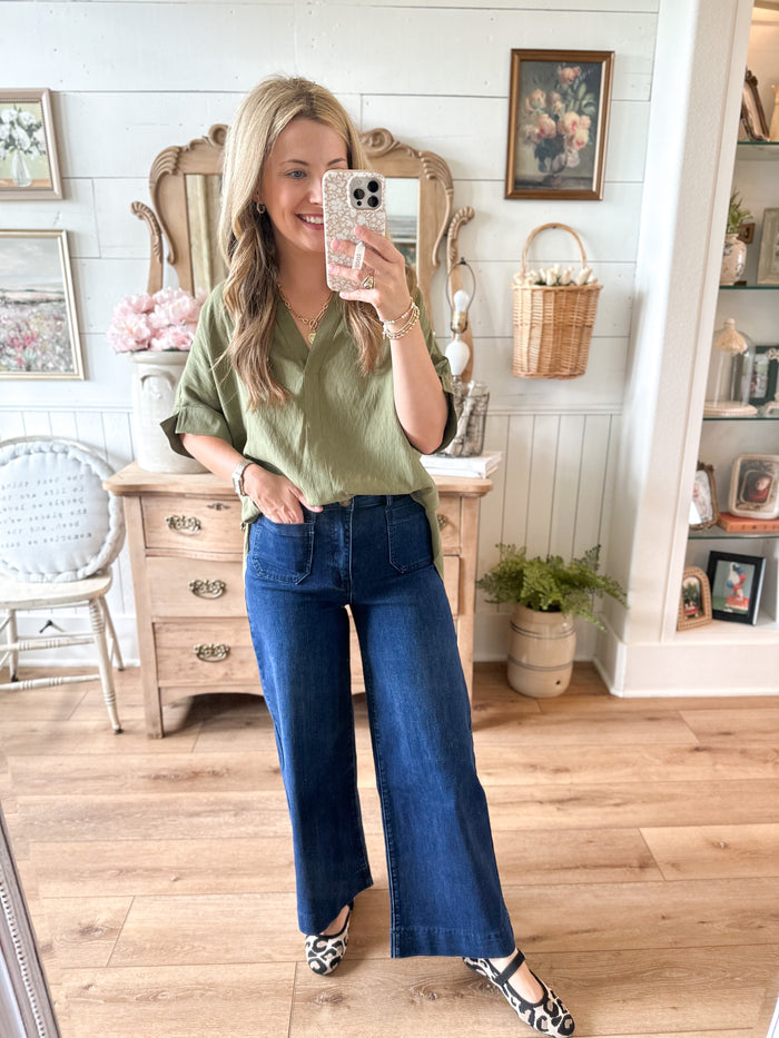 Woman taking a mirror selfie wearing a green shirt and blue jeans in a home setting.