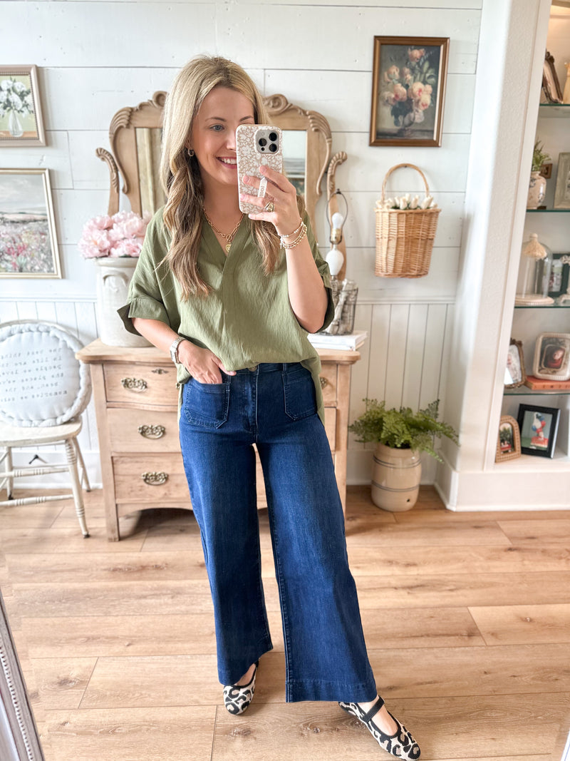 Woman taking a mirror selfie wearing a green shirt and blue jeans in a home setting.