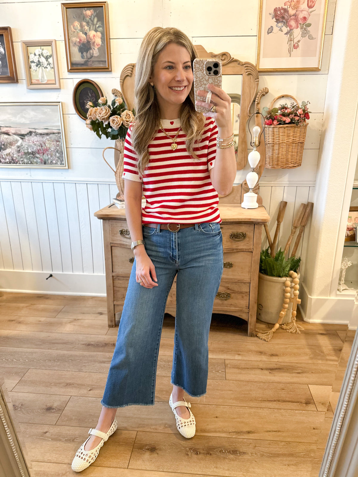 A woman takes a mirror selfie in a cozy, vintage-style room, wearing a red and white striped short-sleeve t-shirt, blue cropped flare jeans, and white Mary Jane shoes.