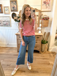 A woman takes a mirror selfie in a cozy, vintage-style room, wearing a red and white striped short-sleeve t-shirt, blue cropped flare jeans, and white Mary Jane shoes.