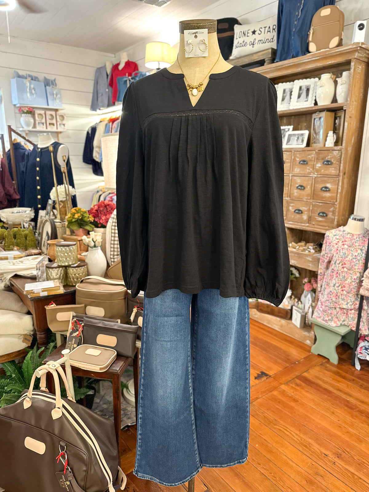 A black long-sleeve blouse with a gathered neckline and puff sleeves, displayed on a mannequin with wide-leg blue jeans in a boutique setting.