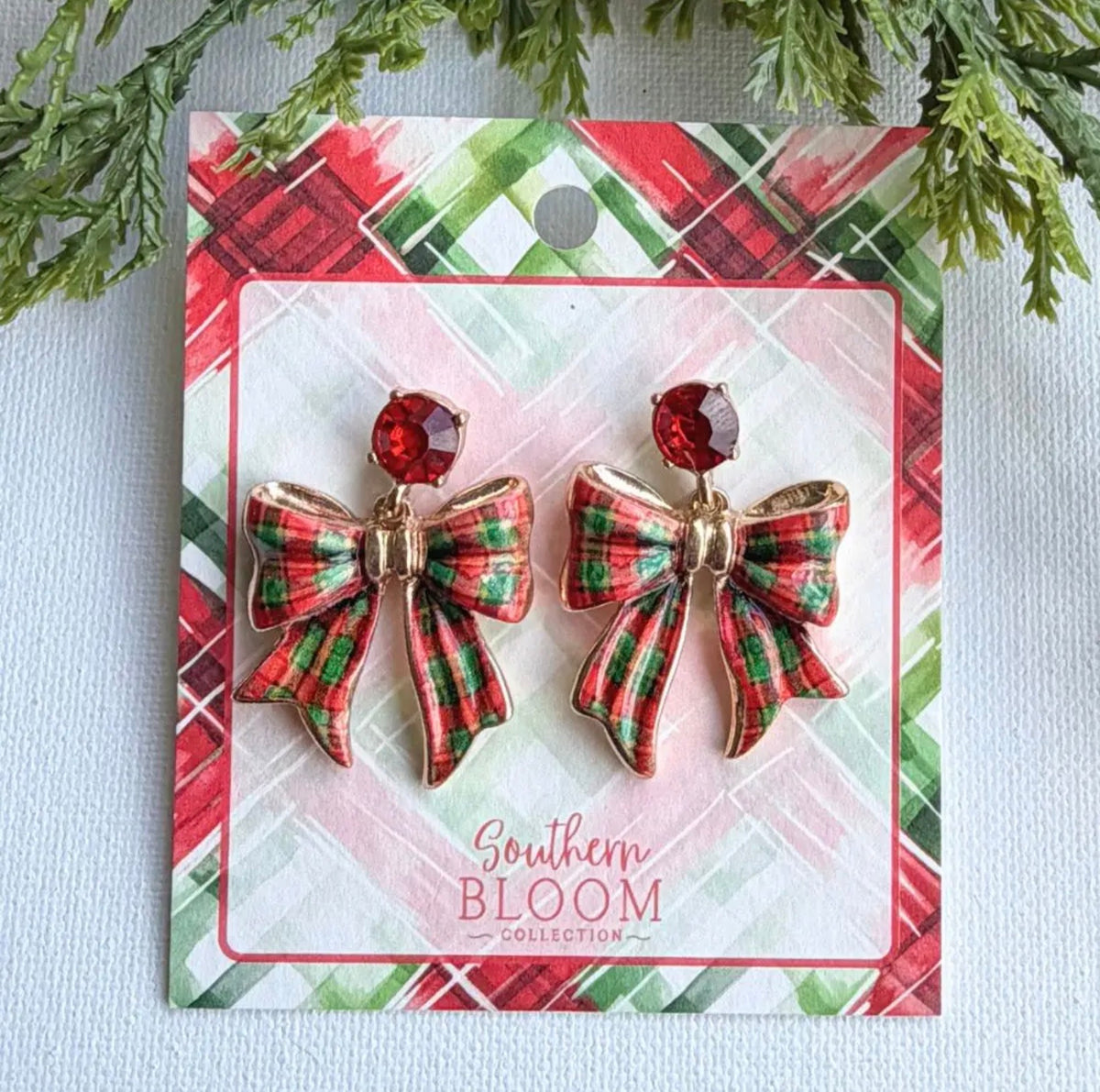 A pair of gold-tone bow earrings with red and green plaid ribbon detailing and a red faceted crystal stud, displayed on a branded Southern Bloom Collection holiday card.