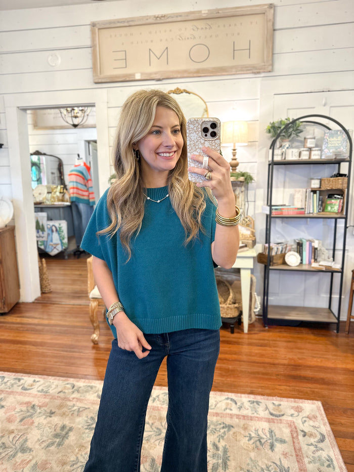 Woman taking a mirror selfie wearing a teal top and dark jeans in a home setting.