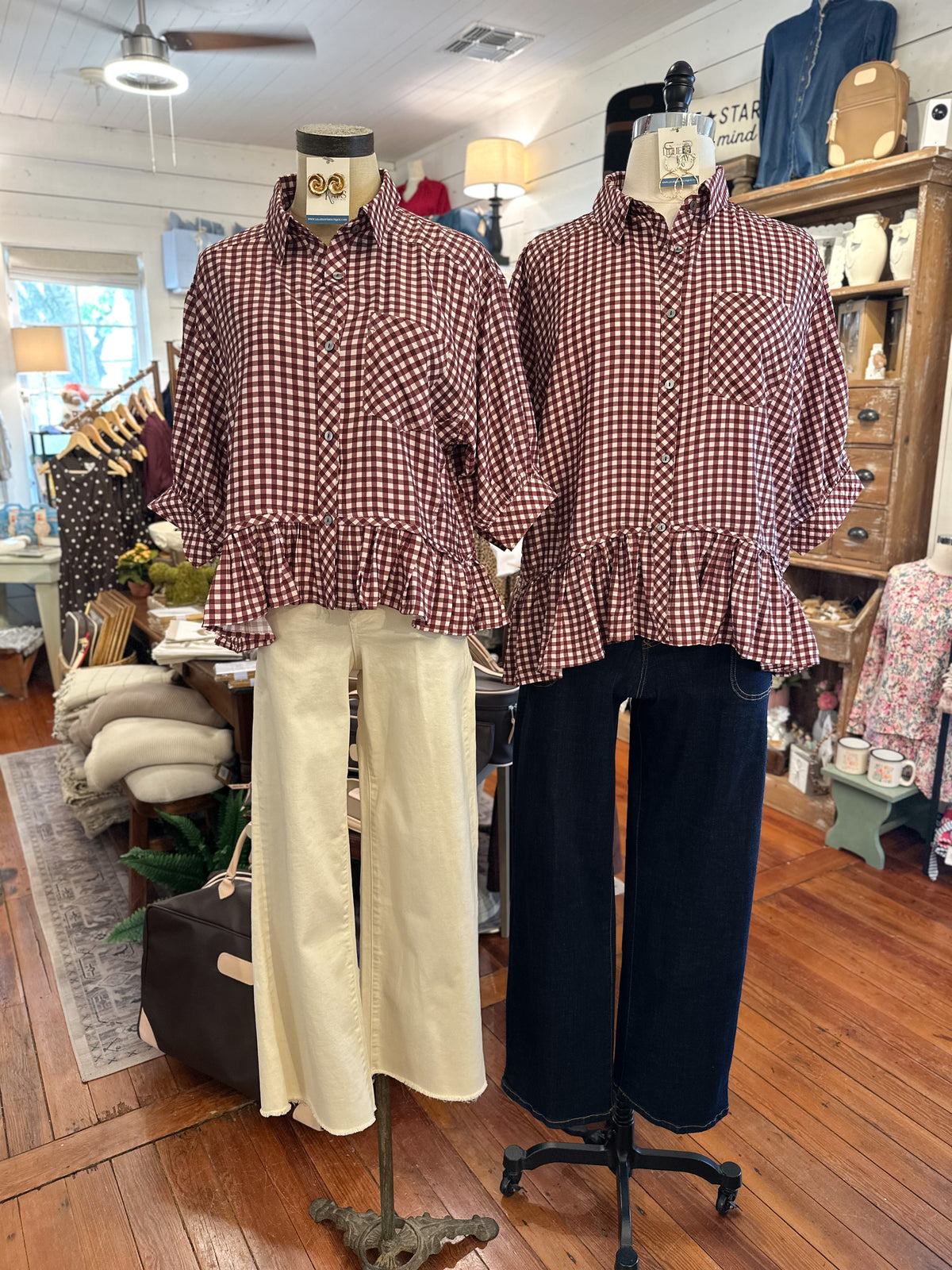 Two women's long-sleeve button-up shirts with a maroon and white gingham check pattern and ruffled peplum hems, displayed on mannequins in a clothing boutique. One is styled with cream wide-leg pants and the other with dark navy wide-leg pants.