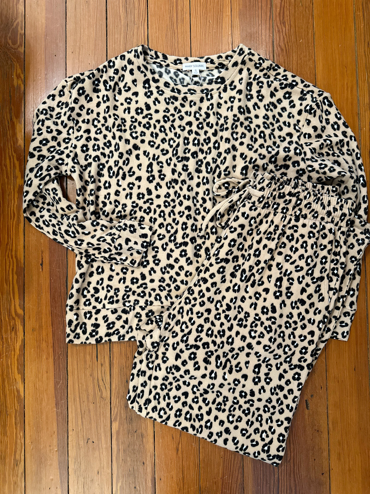 A long-sleeve leopard print top laid flat on a wooden floor. The top has an allover black and dark brown leopard pattern on a warm beige background.
