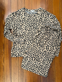 A long-sleeve leopard print top laid flat on a wooden floor. The top has an allover black and dark brown leopard pattern on a warm beige background.