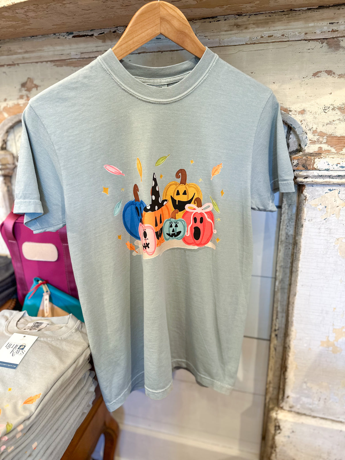 A light sage green short-sleeve t-shirt hanging on a wooden hanger against a rustic distressed wood wall. The shirt features a colorful graphic print of various cartoon pumpkins and ghosts in orange, pink, blue, and green.