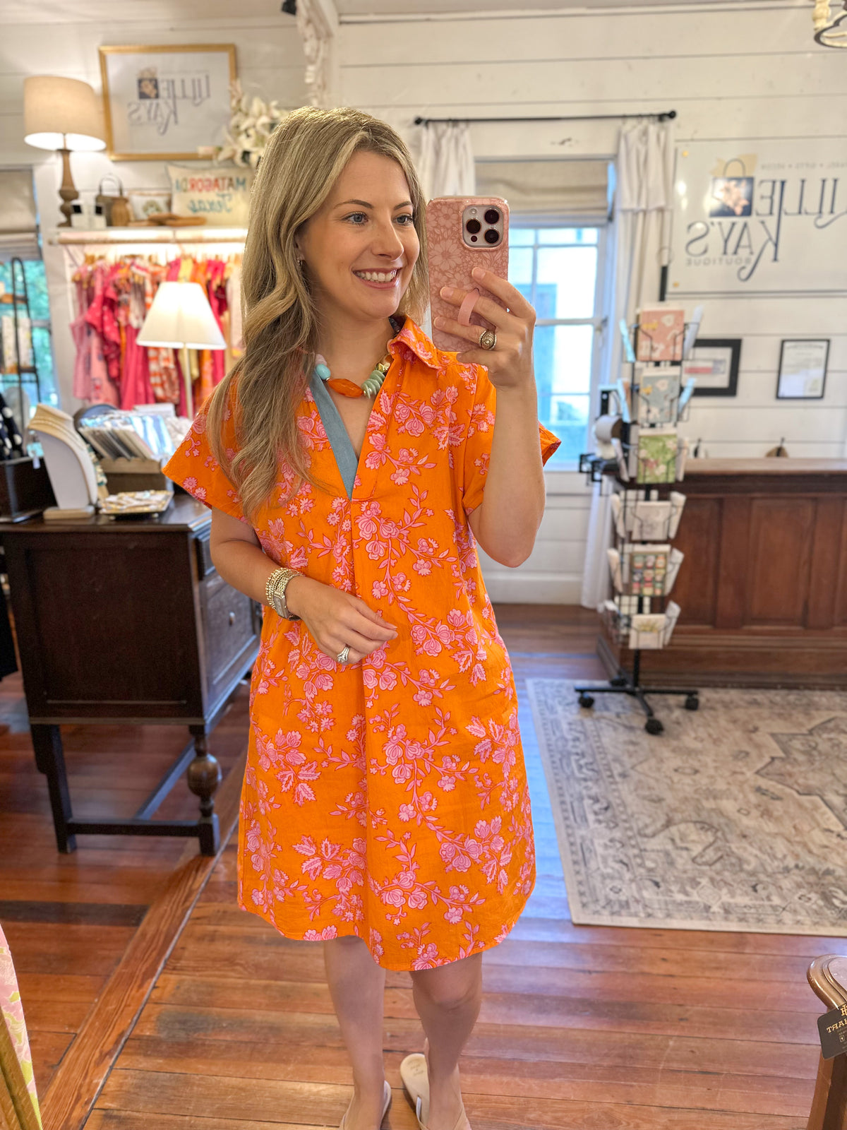 A woman takes a mirror selfie in a boutique shop wearing a bright orange short-sleeve button-up dress with a pink and white floral pattern.