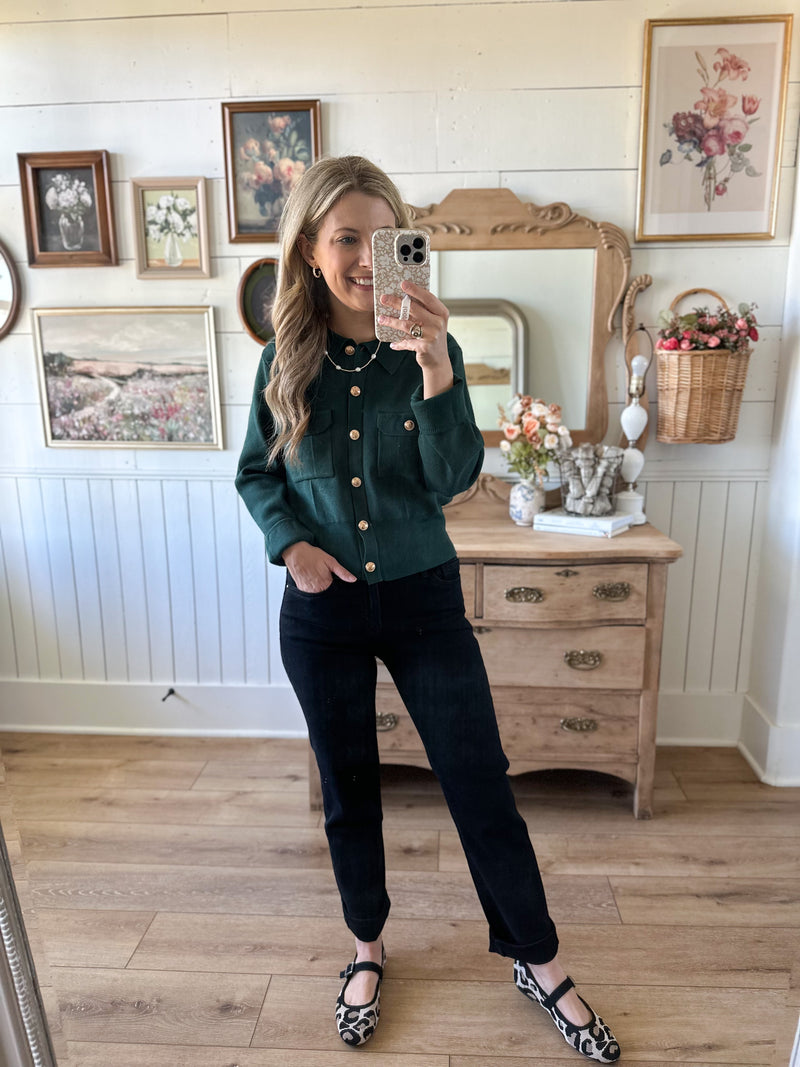 Woman taking a mirror selfie wearing a green jacket and black pants in a room with wooden floor and white walls.