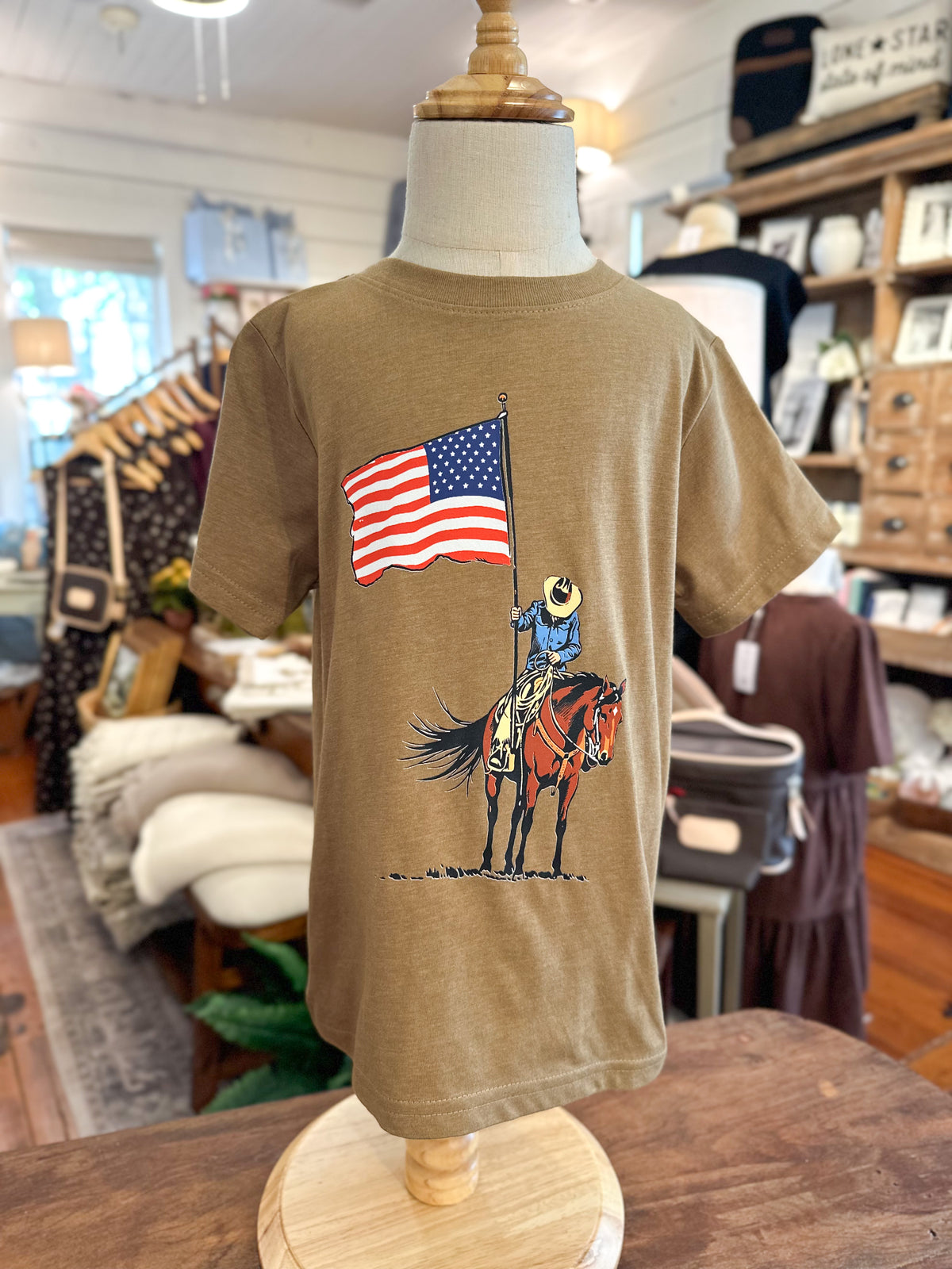 A tan short-sleeve graphic T-shirt displayed on a dress form mannequin inside a retail store. The shirt features a printed graphic of a cowboy on a horse holding a large American flag.